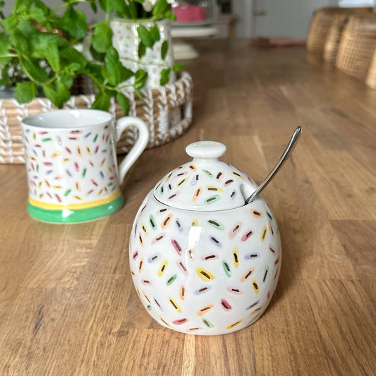 Decorative sugar bowl with colorful sprinkles and a spoon on a wooden table, with a mug and flowers in the background.