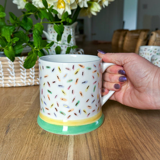 Person holding a Sprinkles XL mug with colorful sprinkles pattern on a wooden table with flowers in the background. Ceramics made by Lianne Peel, Studio LP Ceramics, Ash House studio
