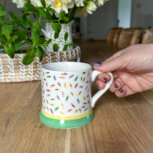 Person holding a sprinkles  mug with colorful patterns on a wooden table with a vase of flowers in the background. Made by Lianne Peel, Studio LP Ceramics, Ash House Studio